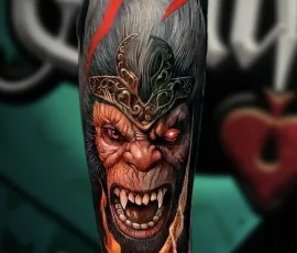 Black and grey realism tattoo of a snarling demon face with red accents, forearm placement